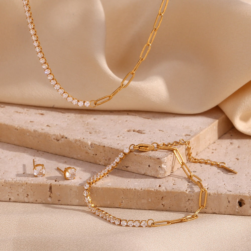 OPUS💎AURI Schmuckset Opus: Wasserfeste 18K PVD-vergoldete Halskette, Armband und Ohrringe im Büroklammer-Design mit Zirkonia, ästhetisch arrangiert auf einer strukturierten Travertin-Natursteinplatte vor einem weich fließenden, beigen Stoffhintergrund.